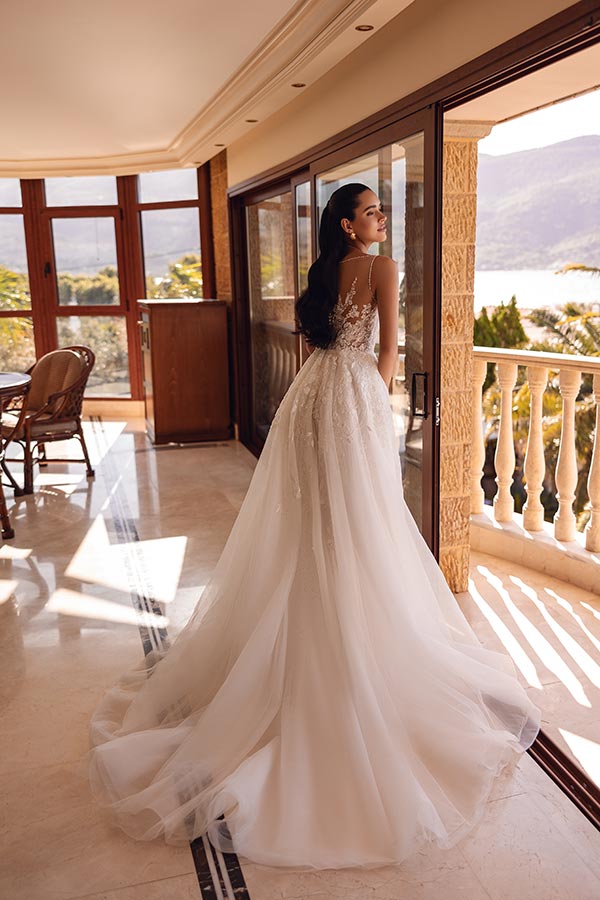 Eine lächelnde Frau im weißen Brautkleid steht in einem sonnigen Raum mit Ausblick