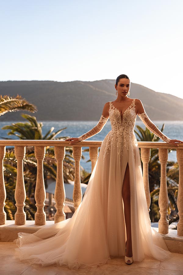 Eine Frau in elegantem cremefarbenem Brautkleid mit Spitzen und Schlitz steht ernst auf einer Terrasse vor Palmen und Meer