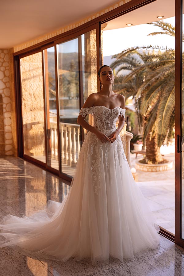 Eine lächelnde Frau im eleganten Brautkleid steht vor großen Fenstern mit Palmenblick