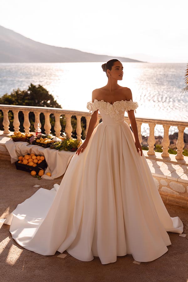 Eine Frau im eleganten weißen Brautkleid steht lächelnd auf einer sonnigen Terrasse am Meer