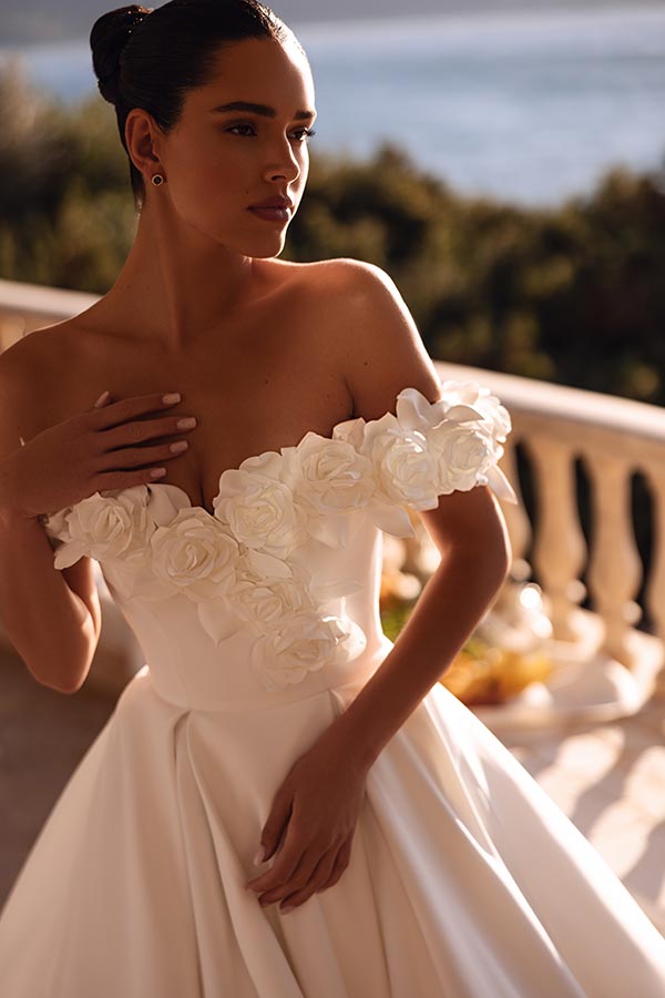 Eine junge Frau mit ernstem Blick trägt ein weißes Kleid mit Blumenapplikationen