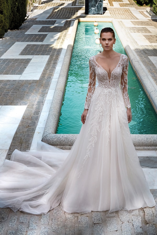 Eine Frau im weißen Brautkleid mit Spitze steht ernst am Rand eines langen Pools