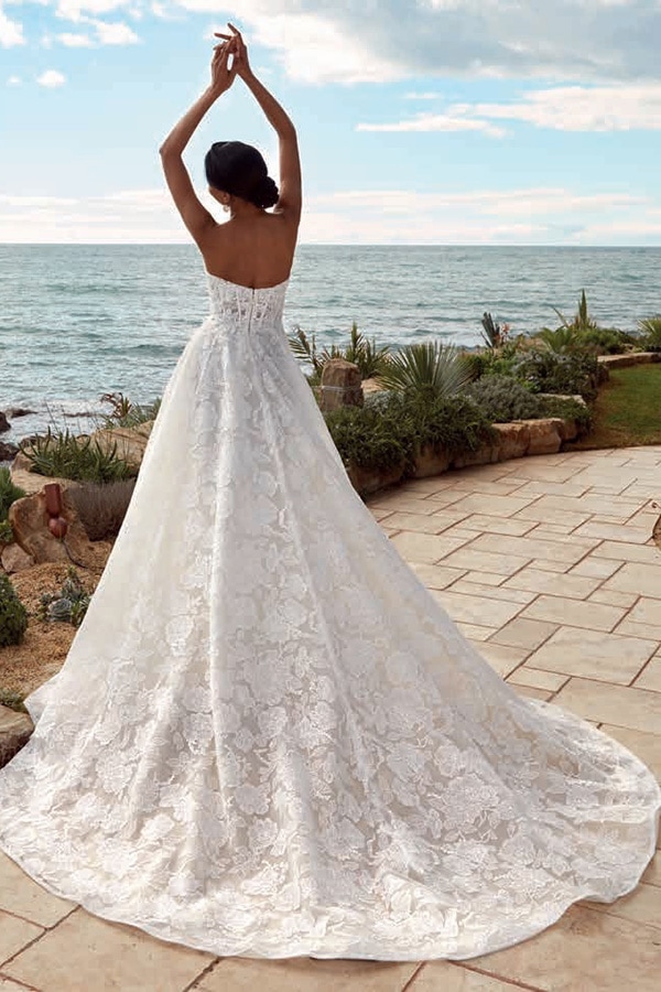 Eine Frau mit Hochsteckfrisur steht im weißen Spitzenkleid am Meer und hebt die Arme