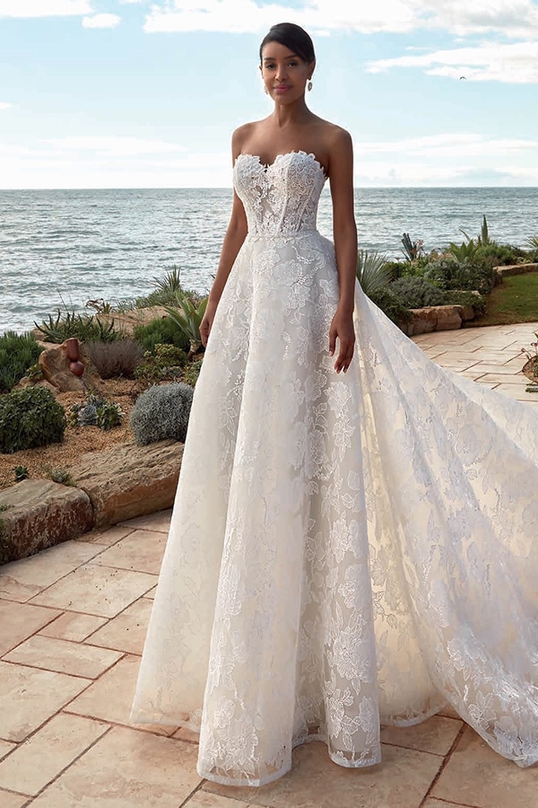 Eine lächelnde Frau in einem weißen Spitzenkleid steht elegant am Meer