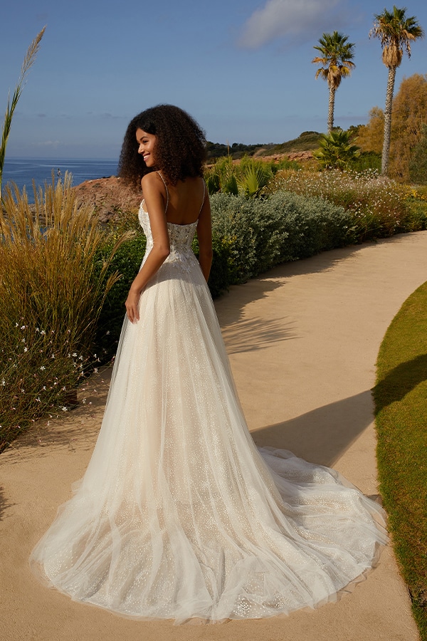 Eine lächelnde Frau mit lockigem Haar trägt ein elegantes weißes Brautkleid am Meer