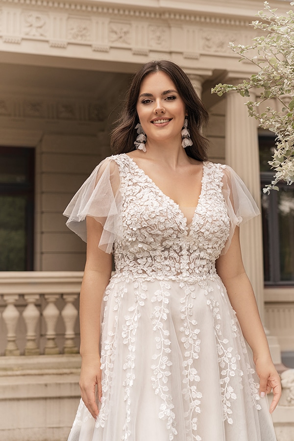 Eine lachende Frau in einem cremefarbenen Kleid mit Blumenmuster steht vor einer Villa