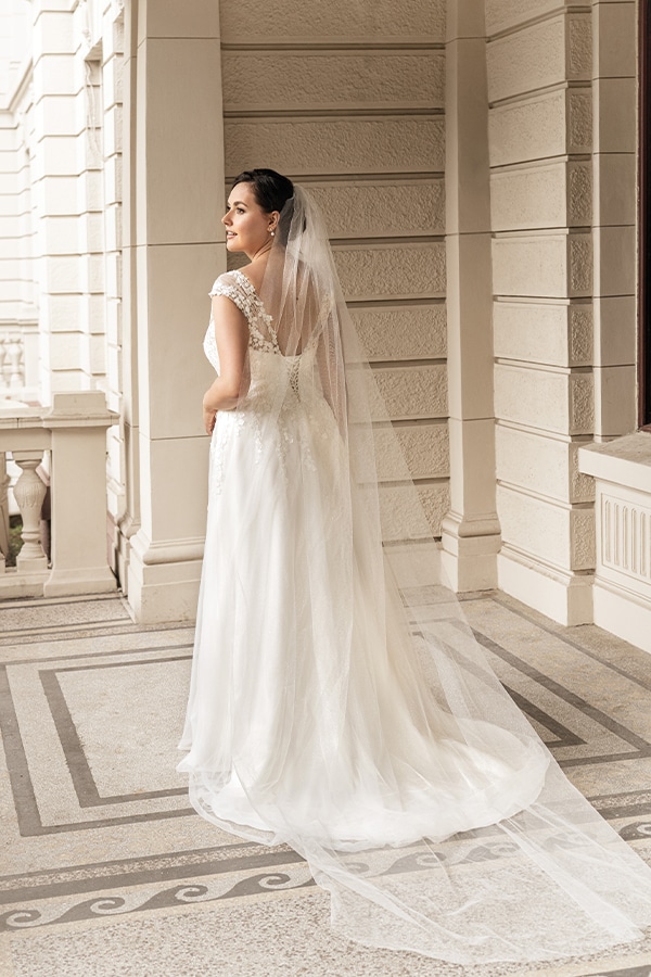 Eine lächelnde Braut im weißen Kleid mit langem Schleier steht auf einer Terrasse