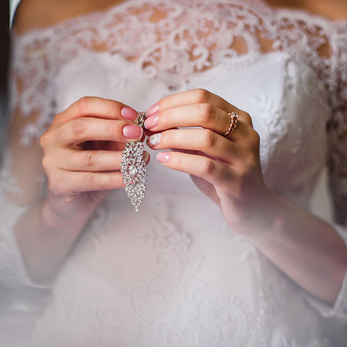 Eine Braut im weißen Spitzenkleid hält funkelnden Ohrschmuck in den Händen