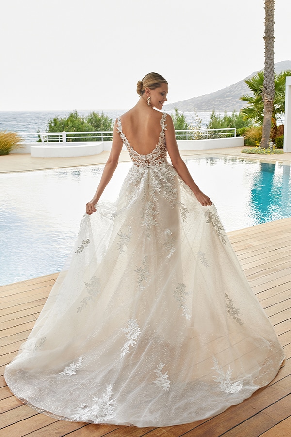 Eine blonde Frau in eleganter Spitzenrobe steht lächelnd am Pool mit Meerblick