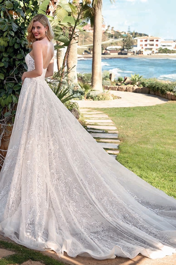 Eine blonde Frau in einem weißen Brautkleid steht lächelnd im Garten am Wasser
