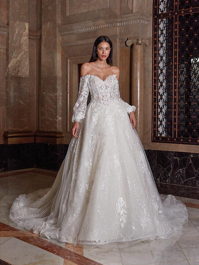 Eine Frau mit langen dunklen Haaren trägt ein elegantes cremefarbenes Brautkleid mit floraler Spitze und blickt ruhig in die Kamera