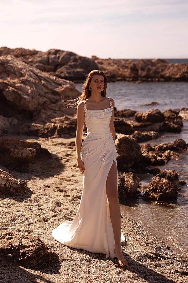 Eine junge Frau mit ernstem Blick trägt ein weißes Kleid am felsigen Meeresufer