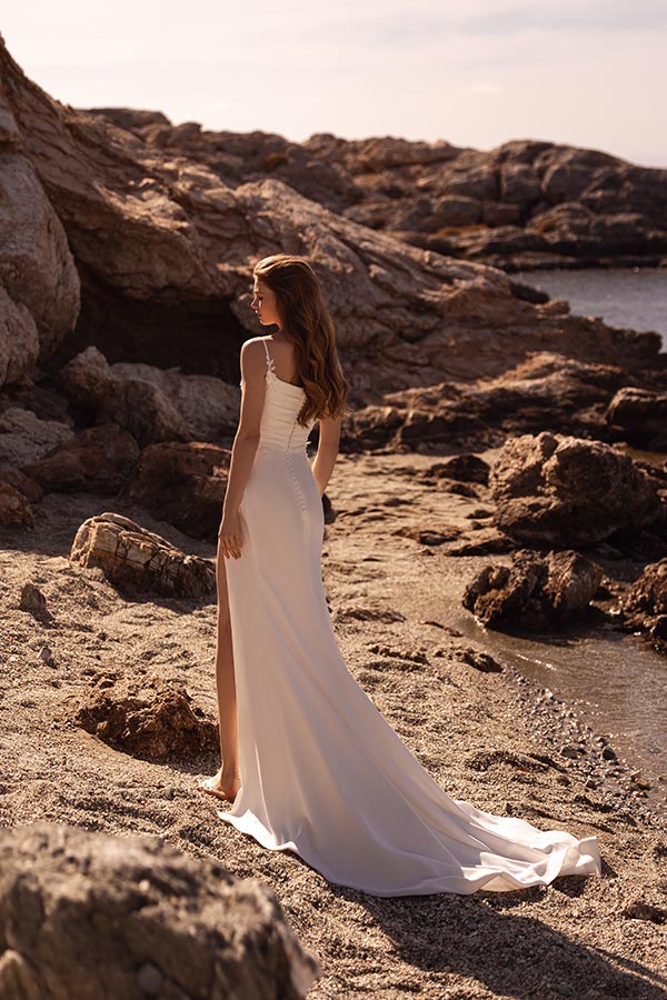 Junge Frau mit langen braunen Haaren im weißen Kleid steht lächelnd am Felsenstrand