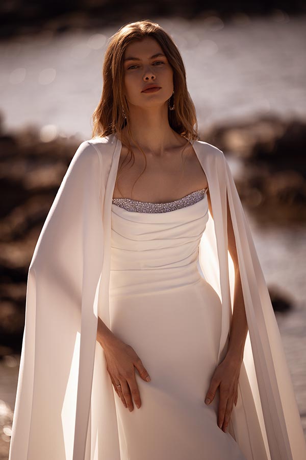 Junge Frau mit langen blonden Haaren trägt elegantes weißes Kleid und capeartiges Tuch am Wasser