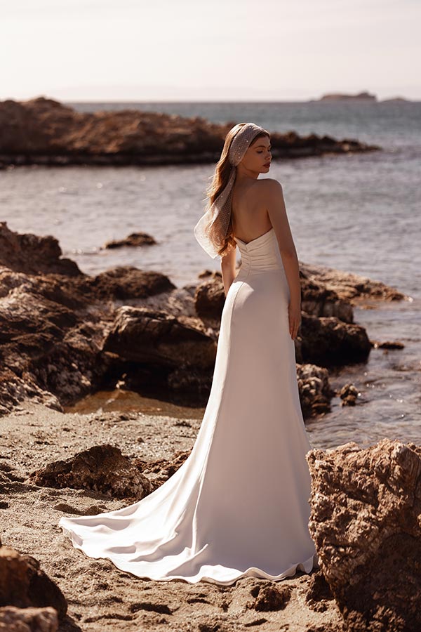 Eine Frau im weißen Brautkleid steht am Felsenstrand und blickt ruhig zum Meer