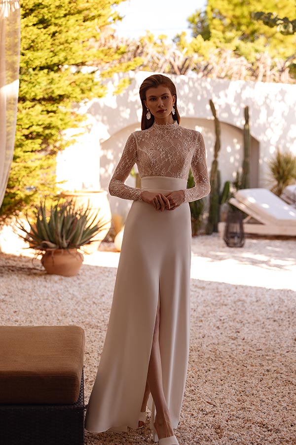 Eine junge Frau mit ernstem Gesichtsausdruck trägt ein elegantes weißes Kleid mit Spitzenärmeln und steht in einem sonnigen Gartenumfeld