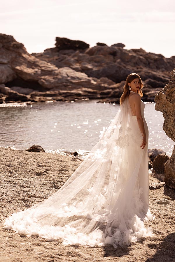 Eine Frau mit langem Schleier steht verträumt am felsigen Strand bei Sonnenlicht