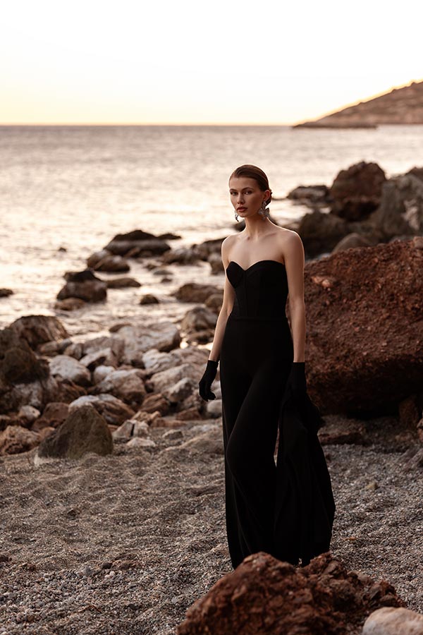 Eine Frau mit ernstem Gesichtsausdruck trägt einen schwarzen Jumpsuit am felsigen Strand