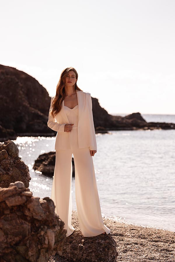 Eine Frau mit langen braunen Haaren steht ernst am Strand im eleganten weißen Hosenanzug
