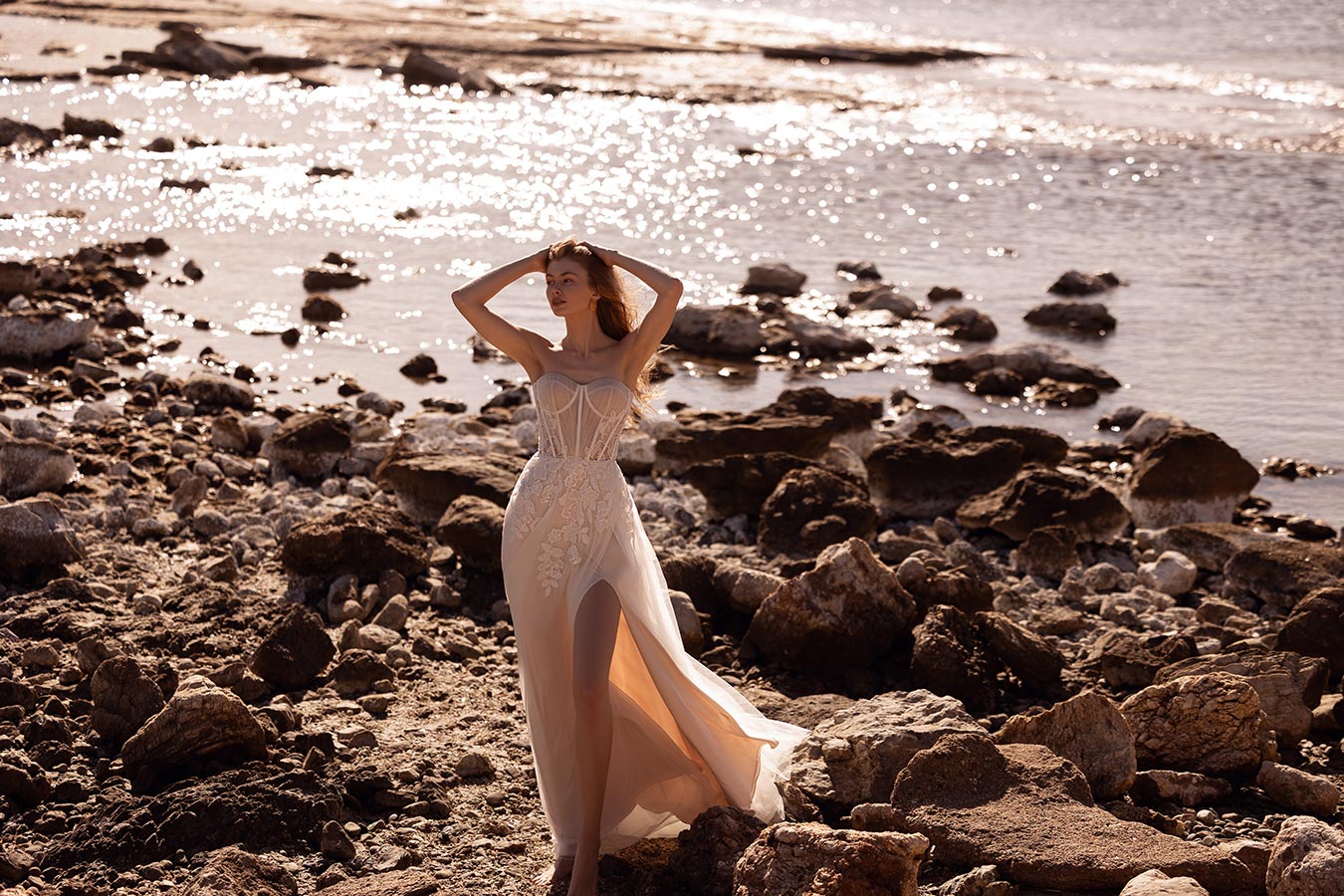Eine Frau mit langem Kleid steht entspannt am felsigen Strand im Sonnenlicht