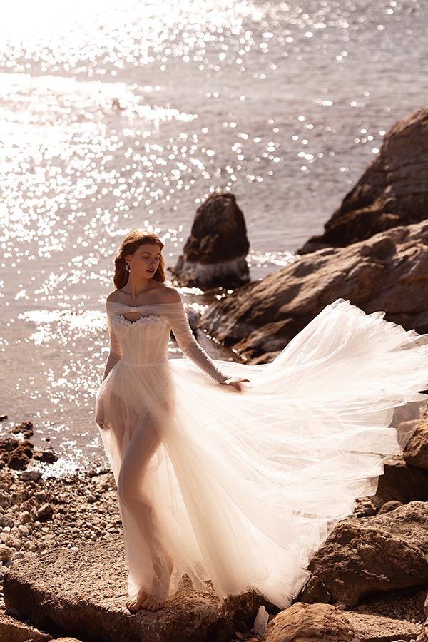 Eine Frau mit sanftem Lächeln trägt ein fließendes weißes Kleid am sonnigen Ufer