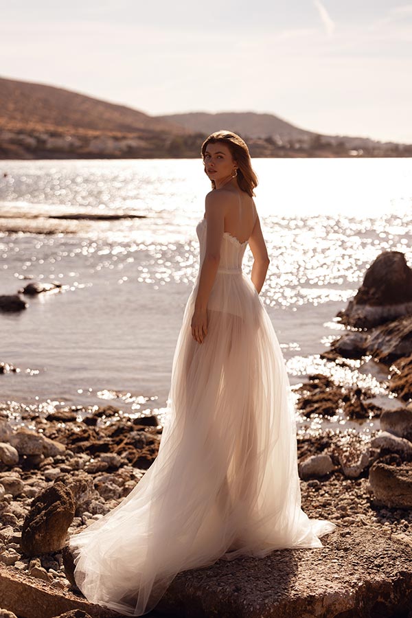 Eine junge Frau mit sanftem Blick steht in einem hellen Kleid am sonnigen Seeufer