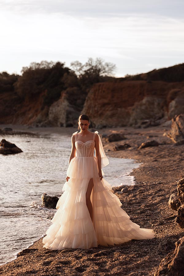 Eine brünette Frau im trägerlosen, weißen Brautkleid steht ernst am Strand