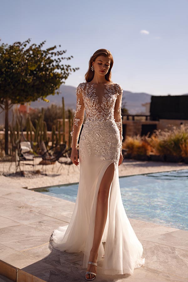 Eine junge Frau mit neutralem Gesichtsausdruck trägt ein elegantes, weißes Kleid mit Spitzenapplikationen und hohem Beinschlitz, während sie am Pool entlanggeht