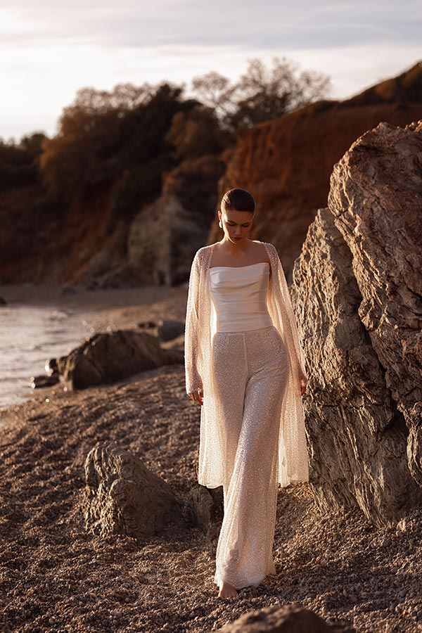 Eine Frau mit neutralem Gesichtsausdruck trägt ein schimmerndes weißes Outfit am Felsenstrand