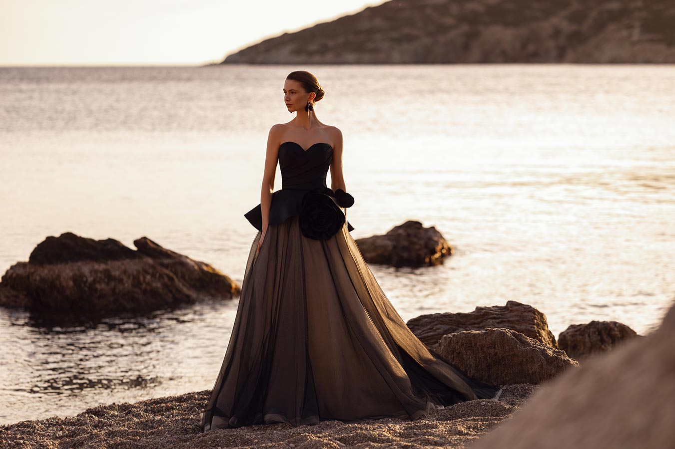 Eine Frau im eleganten schwarzen Kleid steht gelassen am felsigen Meeresufer