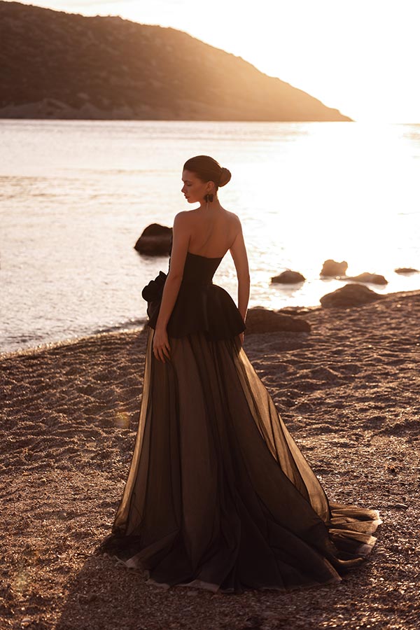 Eine elegant gekleidete Frau in schwarzer Abendrobe steht nachdenklich am Strand im Sonnenuntergang