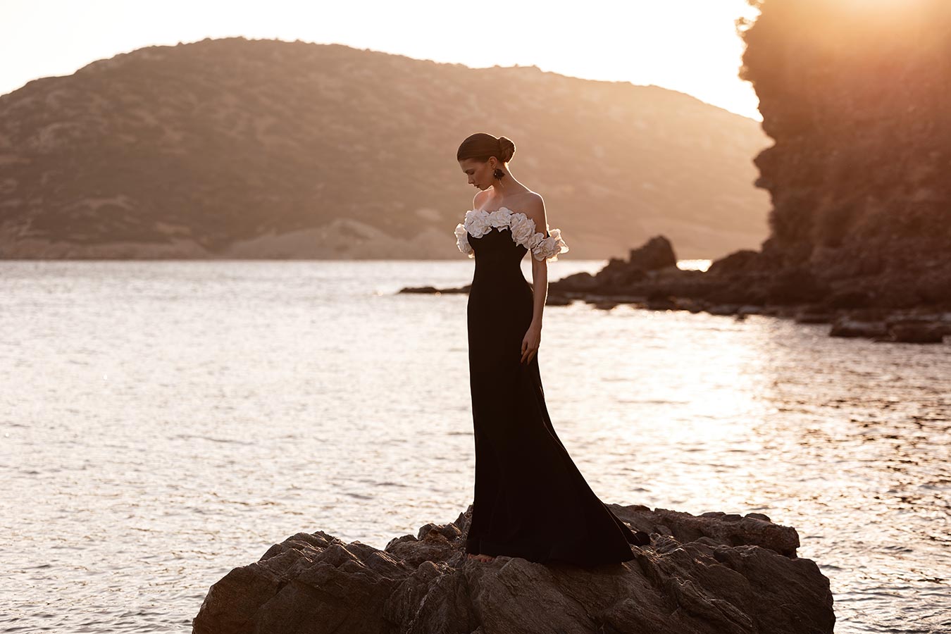 Eine Frau im eleganten schwarzen Kleid mit weißen Blumen steht nachdenklich auf Felsen am Wasser