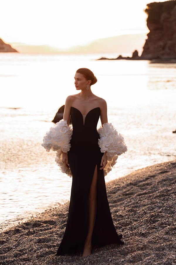 Eine Frau mit elegantem schwarzen Kleid und weißen Blüten steht am Strand bei Sonnenuntergang