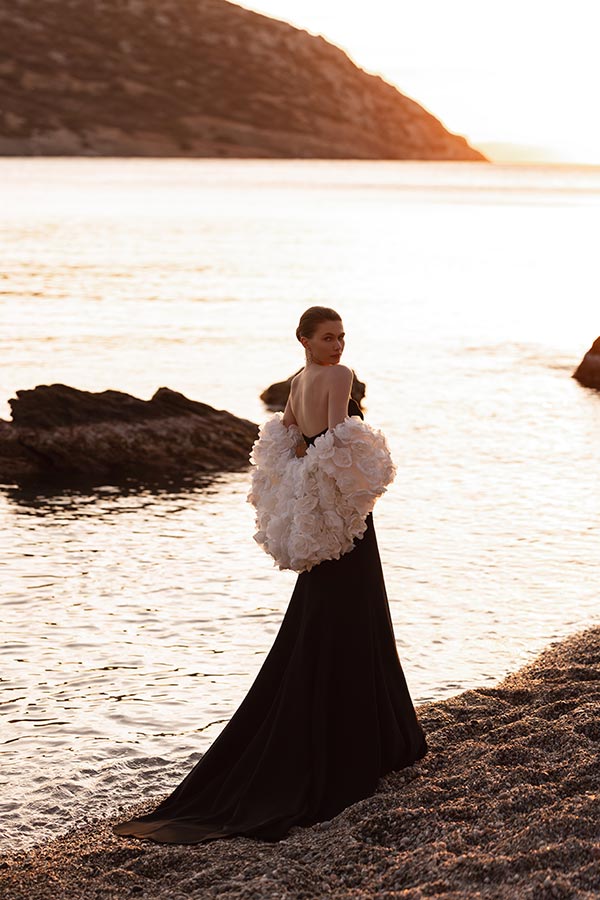 Eine elegant gekleidete Frau in schwarzem Kleid mit weißen Blumenapplikationen steht am Ufer und blickt verträumt über die Schulter