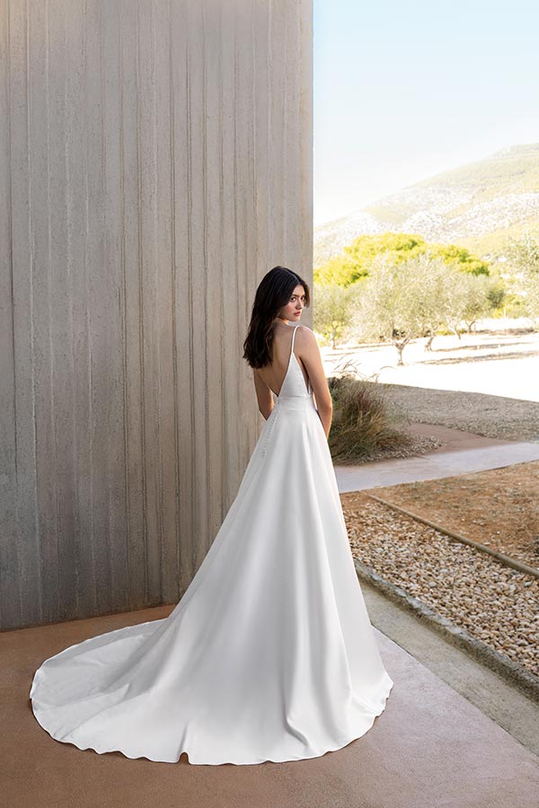 Eine junge Frau mit dunklen Haaren trägt ein elegantes weißes Brautkleid und blickt lächelnd über die Schulter