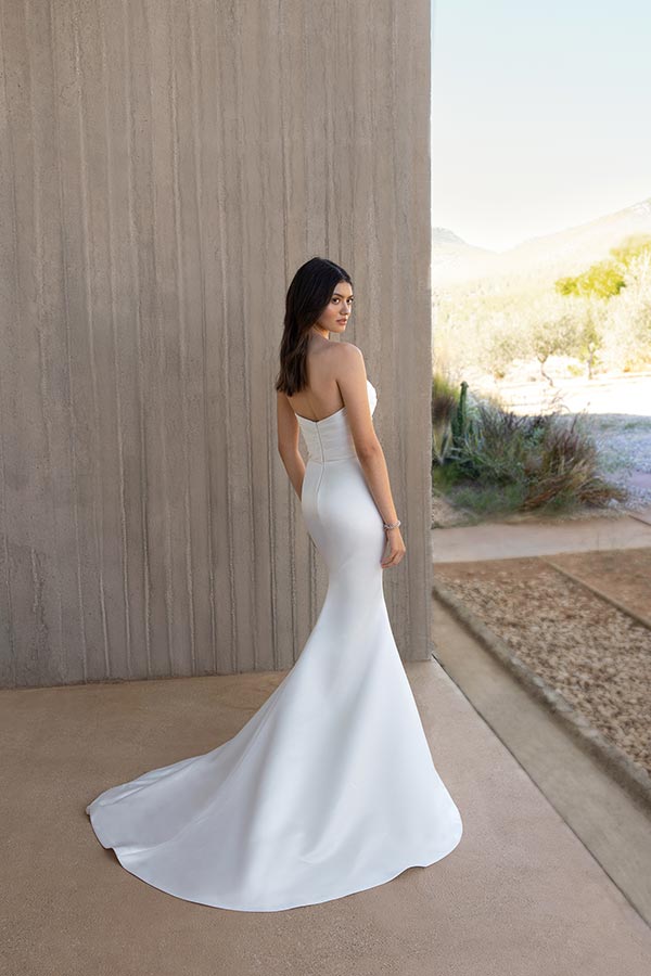 Eine Frau mit langen dunklen Haaren trägt ein elegantes weißes Abendkleid und blickt ruhig über die Schulter in eine sonnige Landschaft