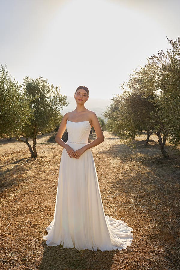 Frau im eleganten weißen Brautkleid steht ernst in sonniger Landschaft mit Bäumen