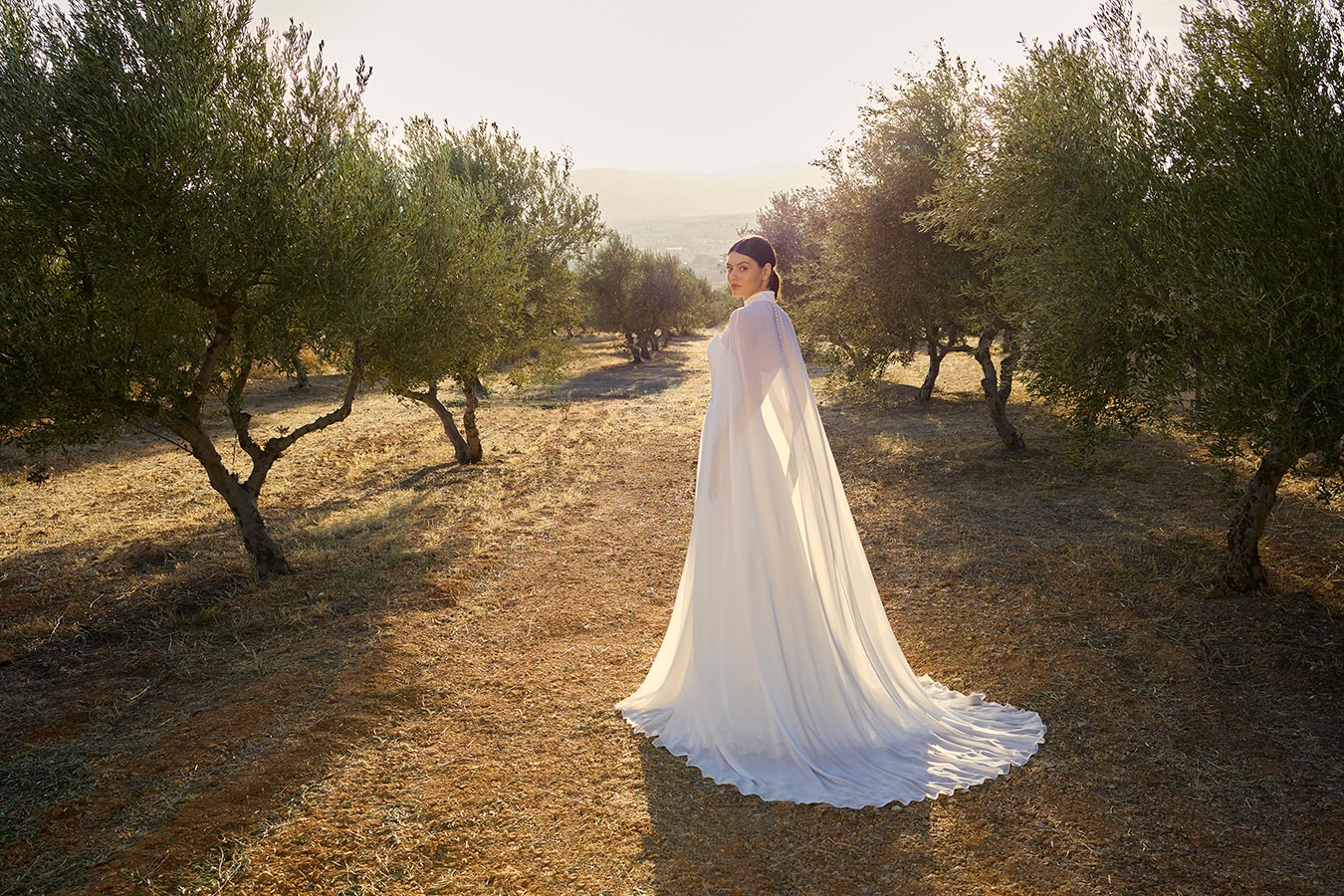 Eine Frau mit hellem Brautkleid steht lächelnd zwischen Olivenbäumen bei Sonnenuntergang