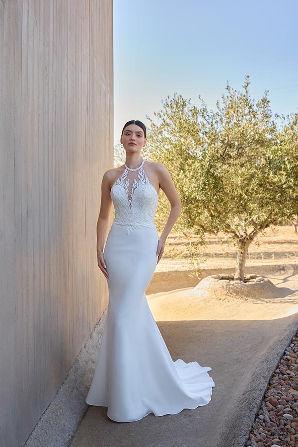 Eine junge Frau mit elegantem weißem Brautkleid steht ernst neben einem Olivenbaum
