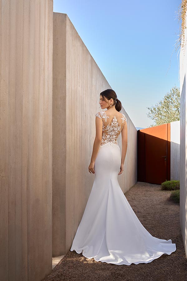 Eine junge Frau im weißen Brautkleid steht lächelnd vor einer Betonwand im Freien