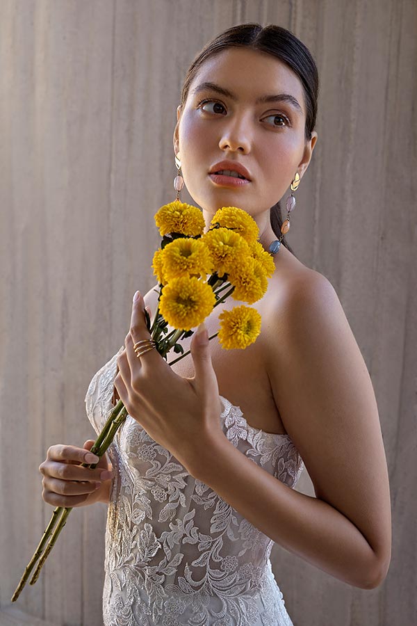 Eine junge Frau mit ernstem Blick hält gelbe Blumen und trägt ein weißes Spitzenkleid