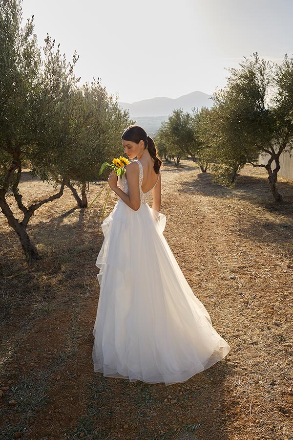 Eine Frau im weißen Brautkleid hält gelbe Blumen und steht nachdenklich im Olivenhain