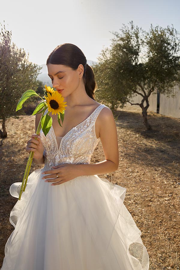 Eine brünette Frau im weißen Brautkleid hält lächelnd eine Sonnenblume und steht im Sonnenschein