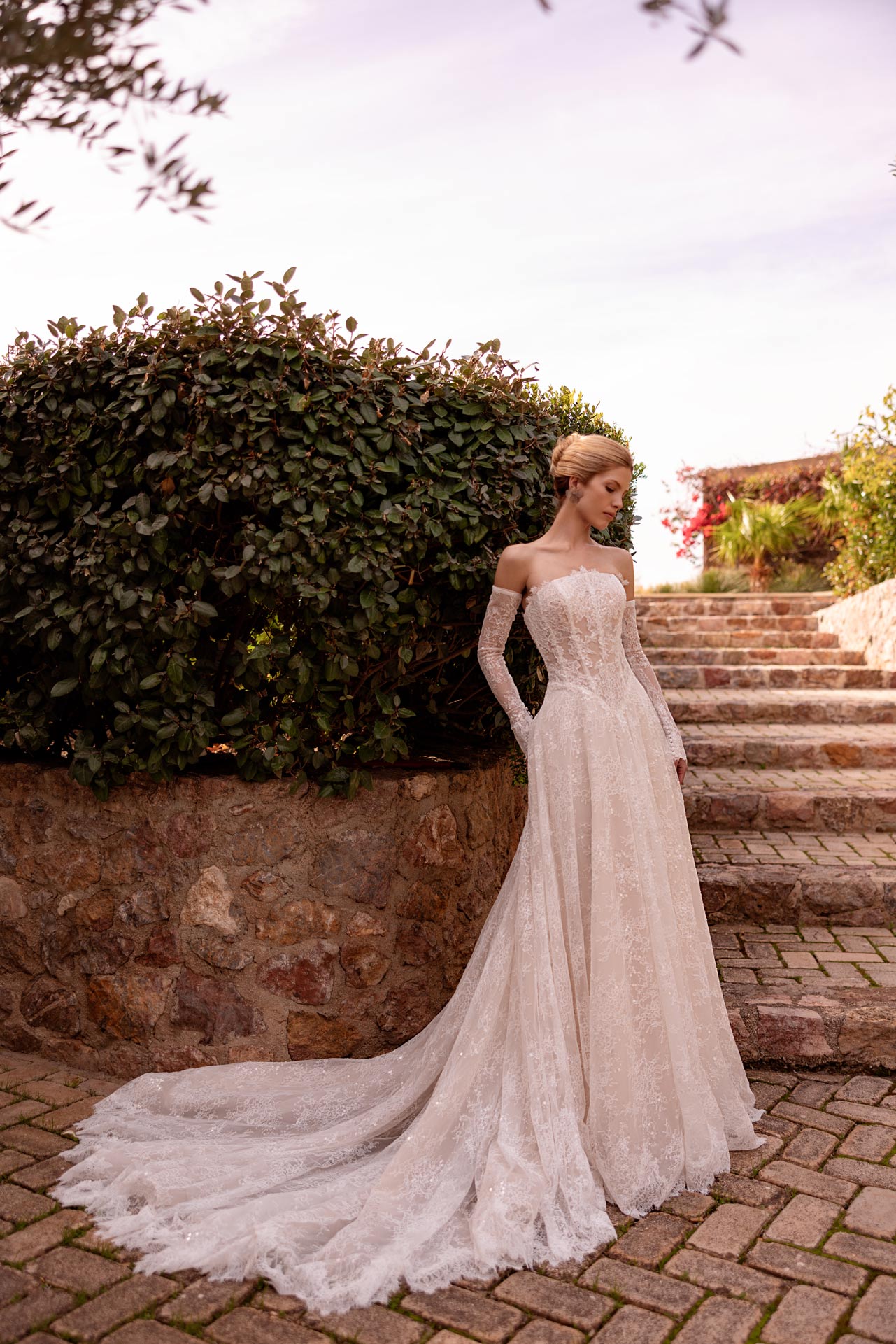 Blonde Braut im eleganten weissen Spitzenkleid mit langer Schleppe im Garten