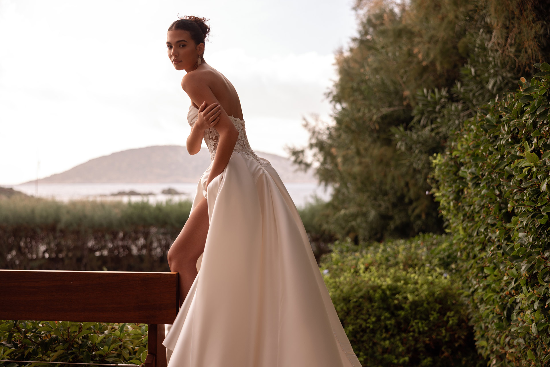 Junge Frau im eleganten weissen Brautkleid mit Beinschlitz vor Kuestenlandschaft