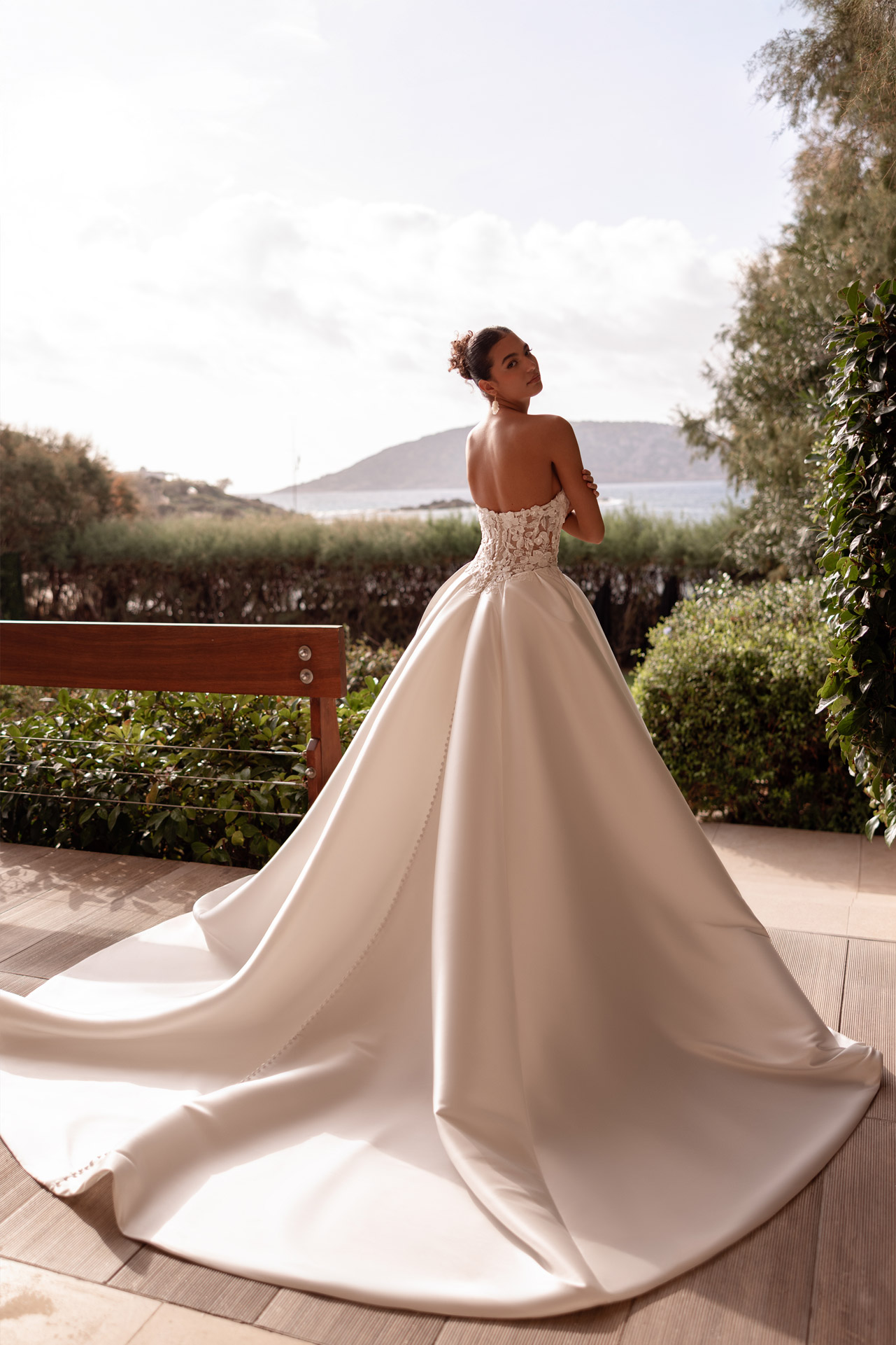 Junge Frau im eleganten weissen Brautkleid mit Schleppe blickt am Meer zurueck