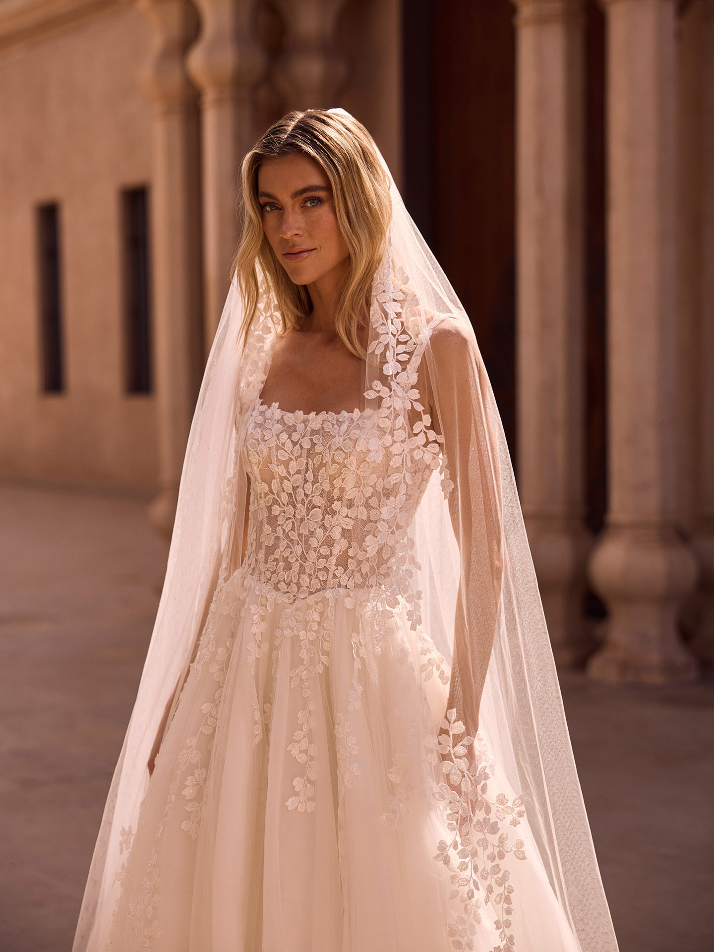 Blonde Braut mit ruhigem Ausdruck im elfenbeinfarbenen Spitzenkleid mit Schleier